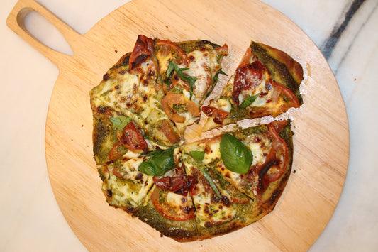 A pesto pizza is served on wooden circular pizza tray. The pizza is cut into slices and made of Besto nut free pesto, mozzarella cheese, prosciutto, basil, and tomato.