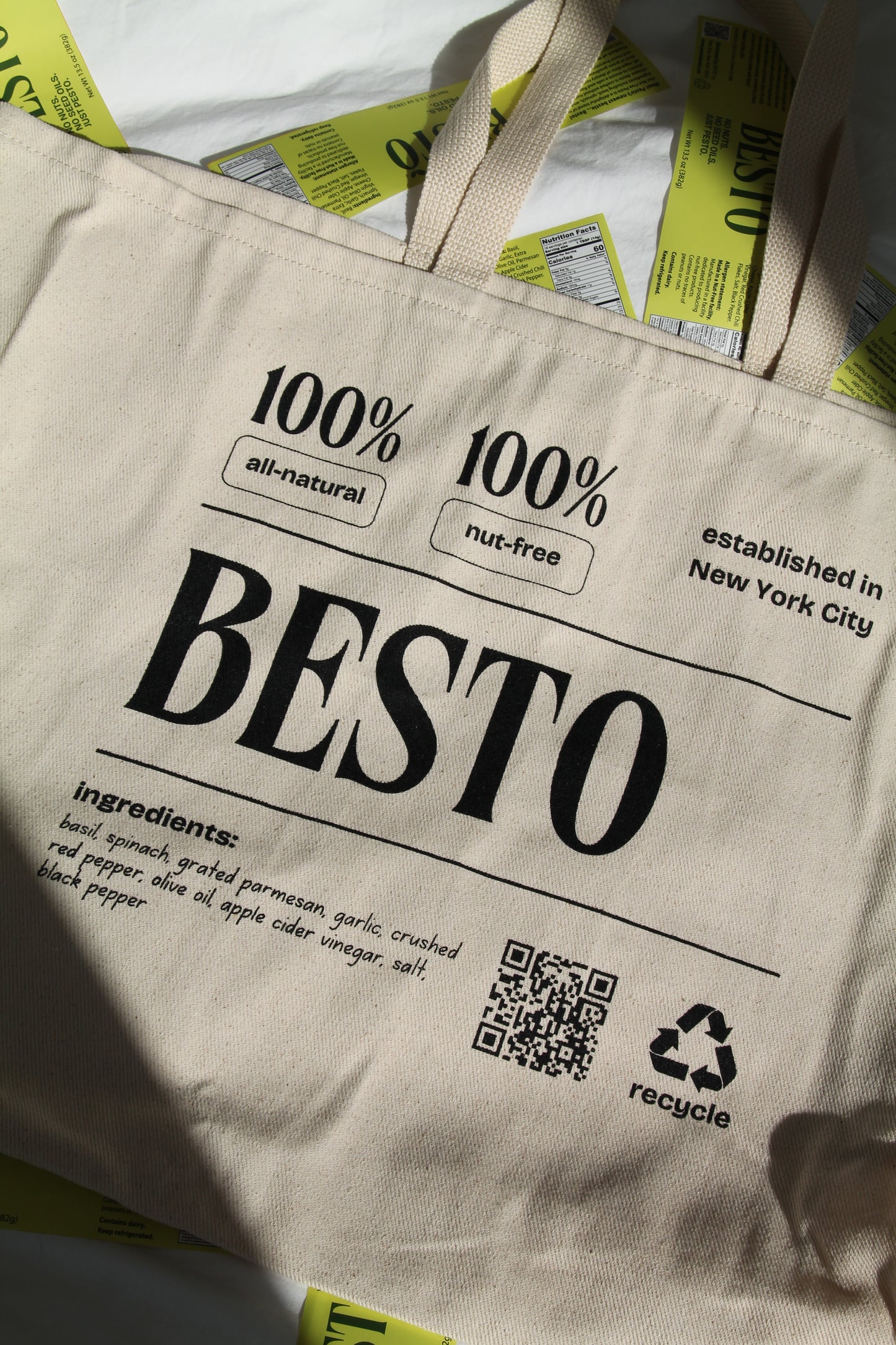 A cream beige large tote bag with "BESTO" in black letters. 100% all natural and nut free. Established in New York City. Recycle. Ingredients listed are basil, spinach, garlic, apple cider vinegar, parmesan cheese, extra virgin olive oil, salt, red crushed chili flakes, and black pepper. QR code.