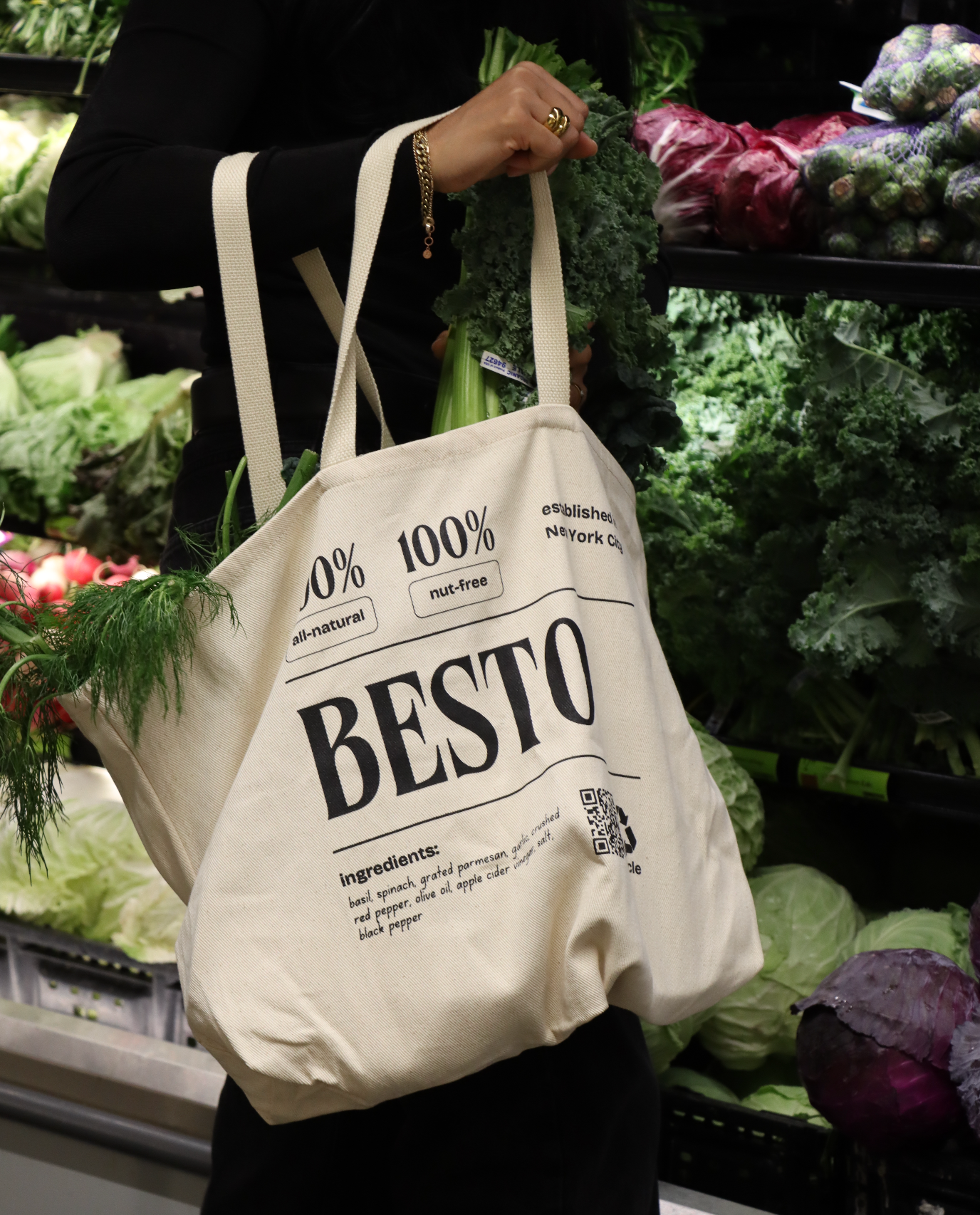 A person carrying a beige, cream tote bag labeled BESTO and 100% all-natural, 100% nut-free while shopping for vegetables. The bag contains green leafy vegetables, and the background shows various fresh produce in a store.