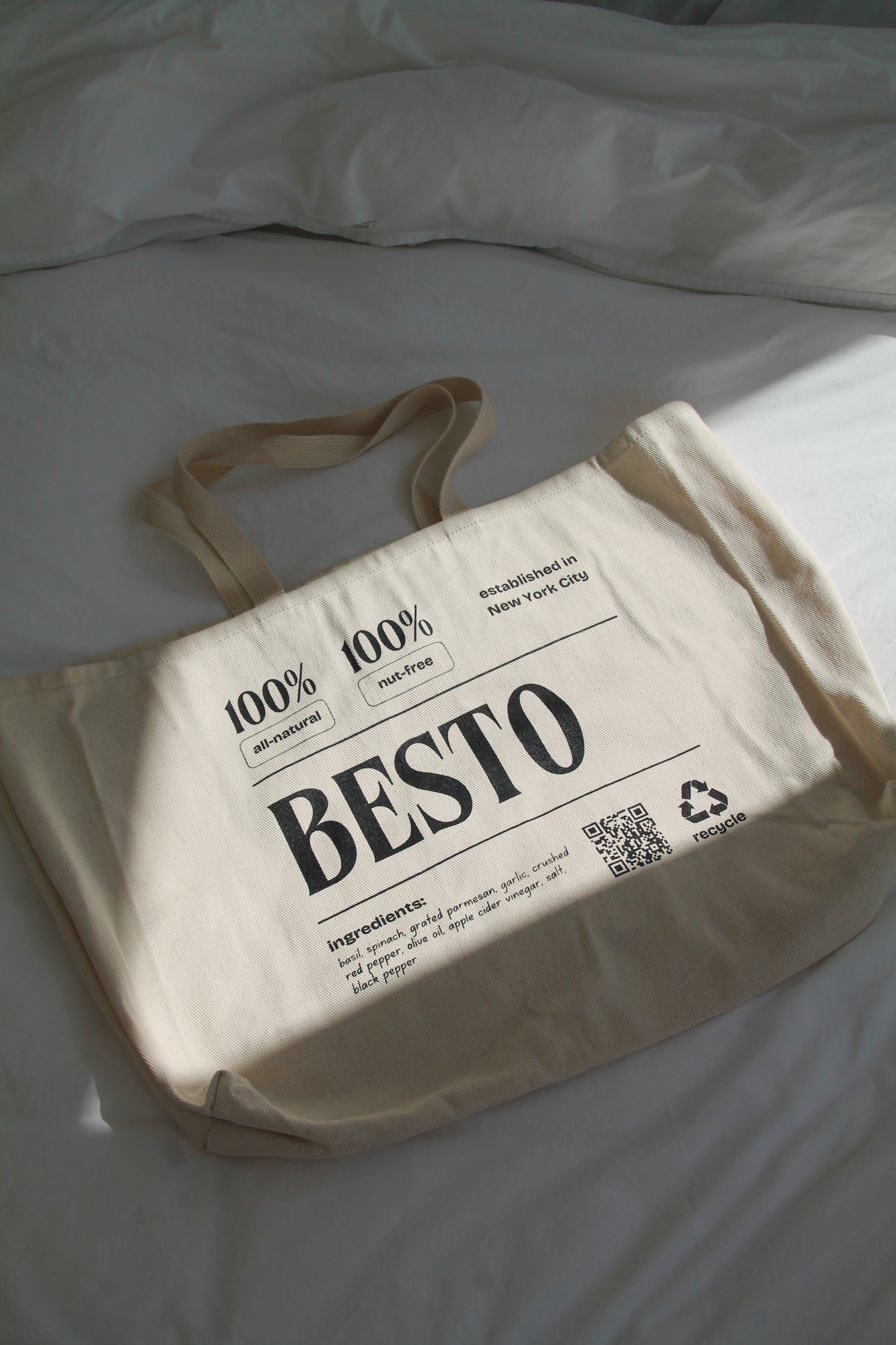 A cream beige large tote bag with "BESTO" in black letters. 100% all natural and nut free. Established in New York City. Recycle. Ingredients listed are basil, spinach, garlic, apple cider vinegar, parmesan cheese, extra virgin olive oil, salt, red crushed chili flakes, and black pepper. QR code.