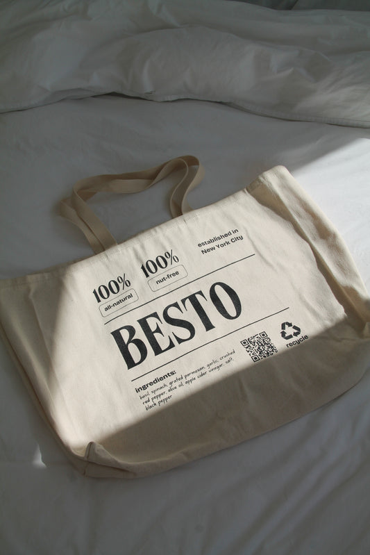 A cream beige large tote bag with "BESTO" in black letters. 100% all natural and nut free. Established in New York City. Recycle. Ingredients listed are basil, spinach, garlic, apple cider vinegar, parmesan cheese, extra virgin olive oil, salt, red crushed chili flakes, and black pepper. QR code.