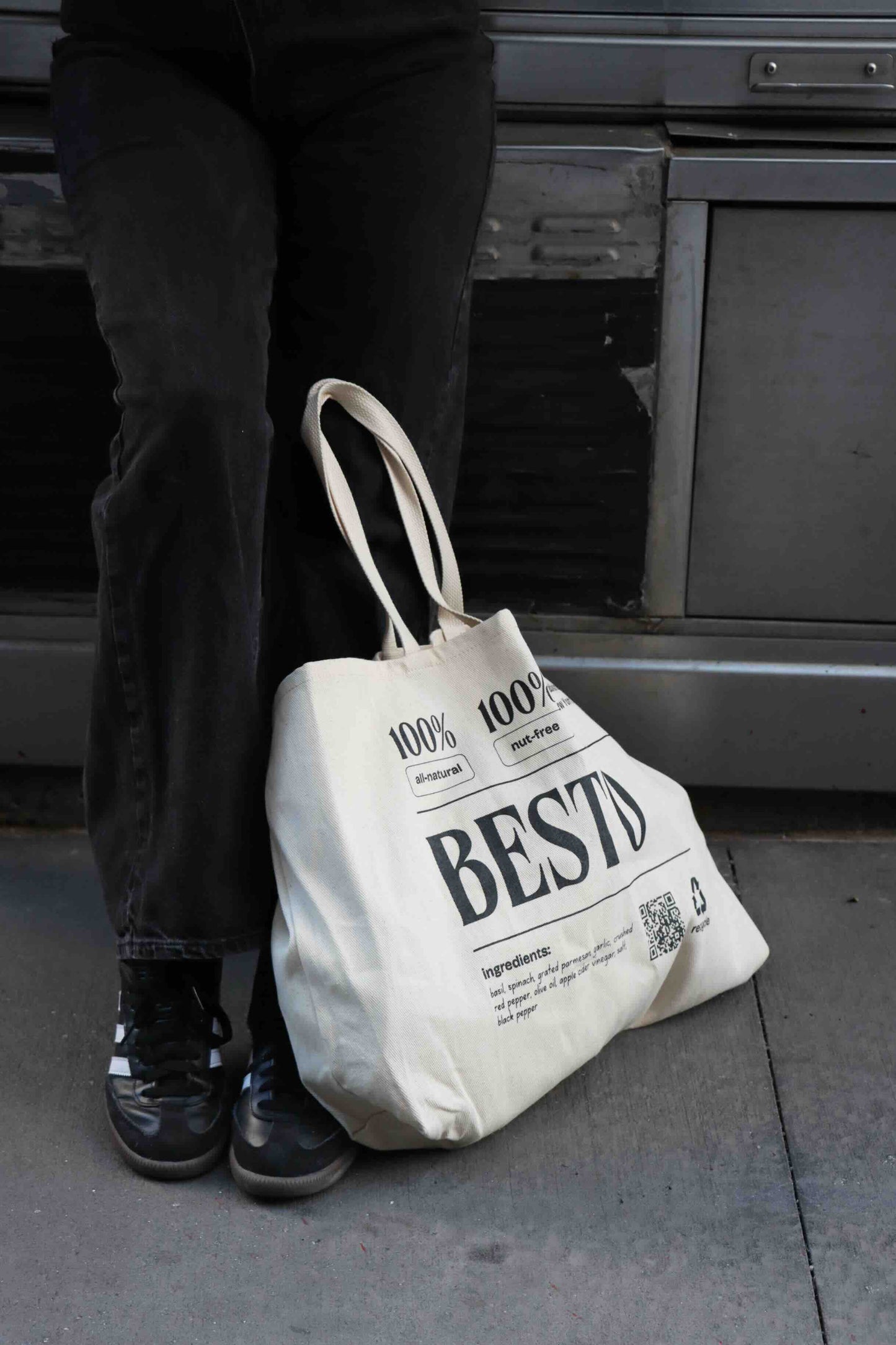 A woman in black jeans and black and white Adidas sambas on a concrete sidewalk. Resting against their leg is a cream beige large tote bag with "BESTO" in black letters. 100% all natural and nut free. Established in New York City. Recycle. Ingredients listed are basil, spinach, garlic, apple cider vinegar, parmesan cheese, extra virgin olive oil, salt, red crushed chili flakes, and black pepper. QR code.