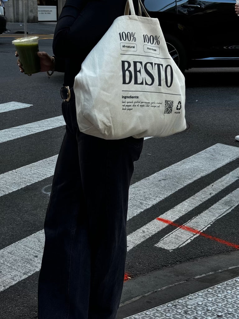 A woman in black jeans and black top holding a green juice. She is standing on a crosswalk in New York City holding a cream beige large tote bag with "BESTO" in black letters. 100% all natural and nut free. Established in New York City. Recycle. Ingredients listed are basil, spinach, garlic, apple cider vinegar, parmesan cheese, extra virgin olive oil, salt, red crushed chili flakes, and black pepper. QR code.