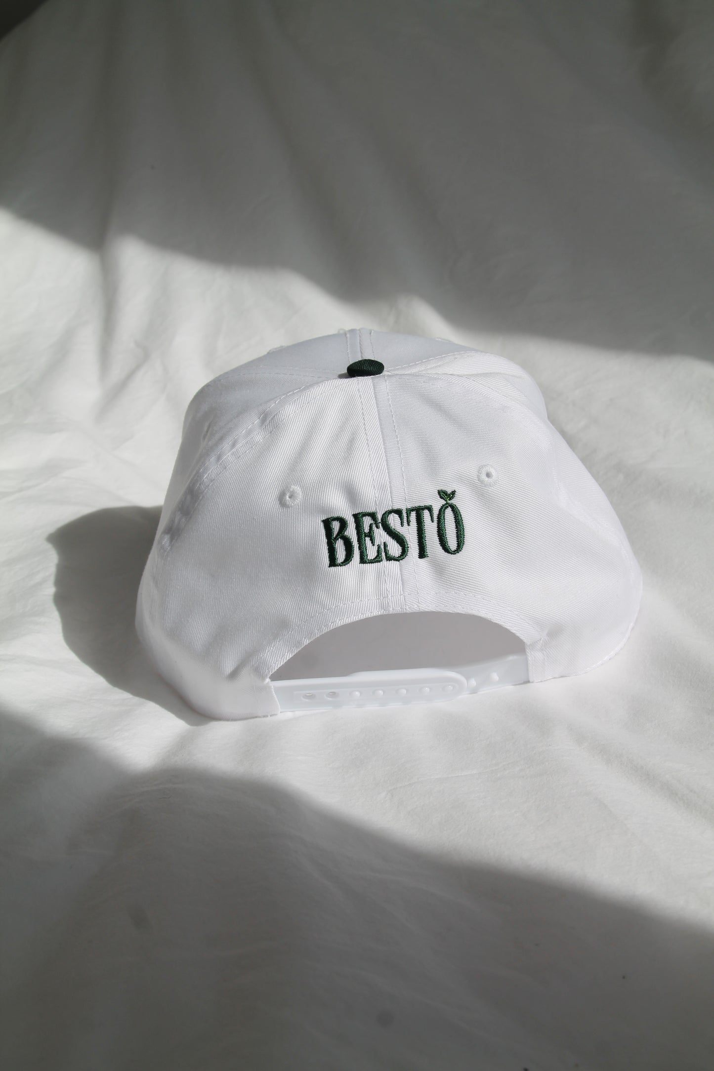 The back of a white trucker hat with dark green letters with the logo "BESTO" with a leaf on the "O". The white strap is adjustable. The hat is sitting on clean, white comforter with natural lighting and shadows.