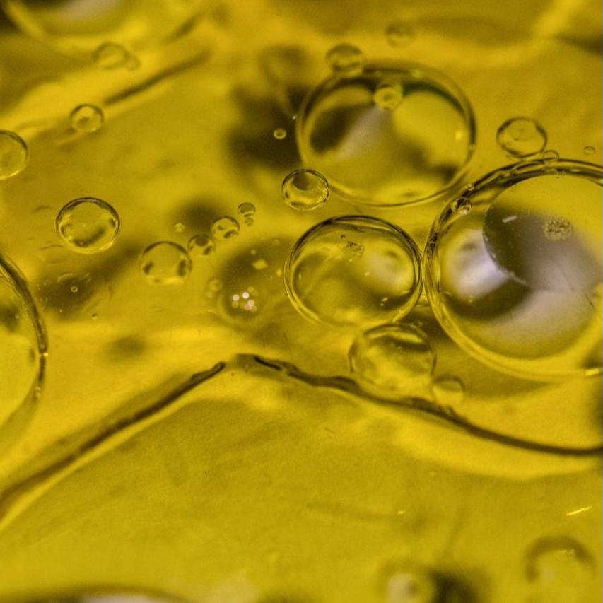 Close-up of oil droplets suspended in yellow liquid, creating a mix of large and small spheres. The background is a uniform yellow, highlighting the transparency and curvature of the droplets.