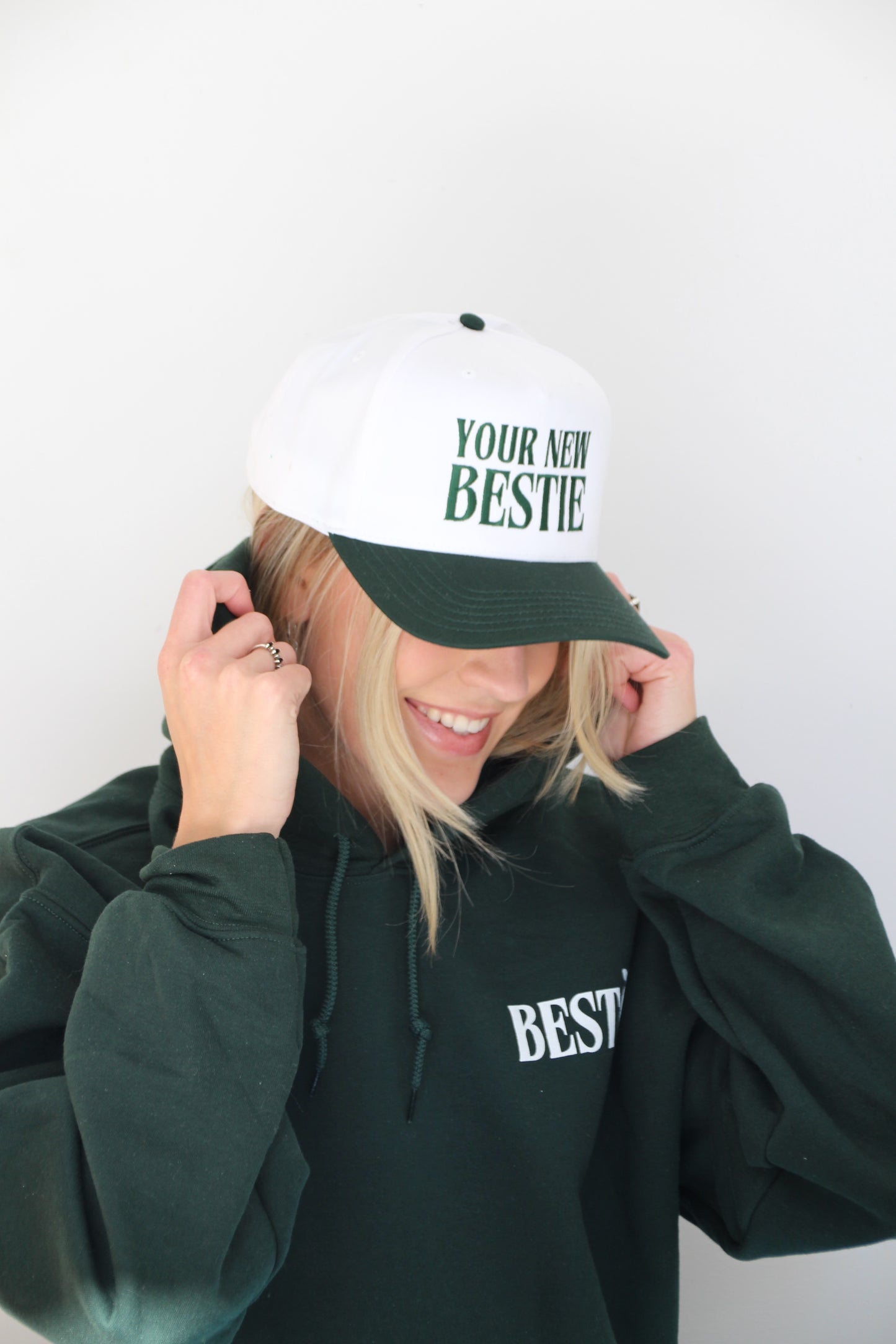 A blonde woman is wearing a white trucker hat with dark green letters embroidered stating "YOUR NEW BESTIE" with a dark green lid. She is smiling and wearing a forest green hoodie with a white logo "BESTO".