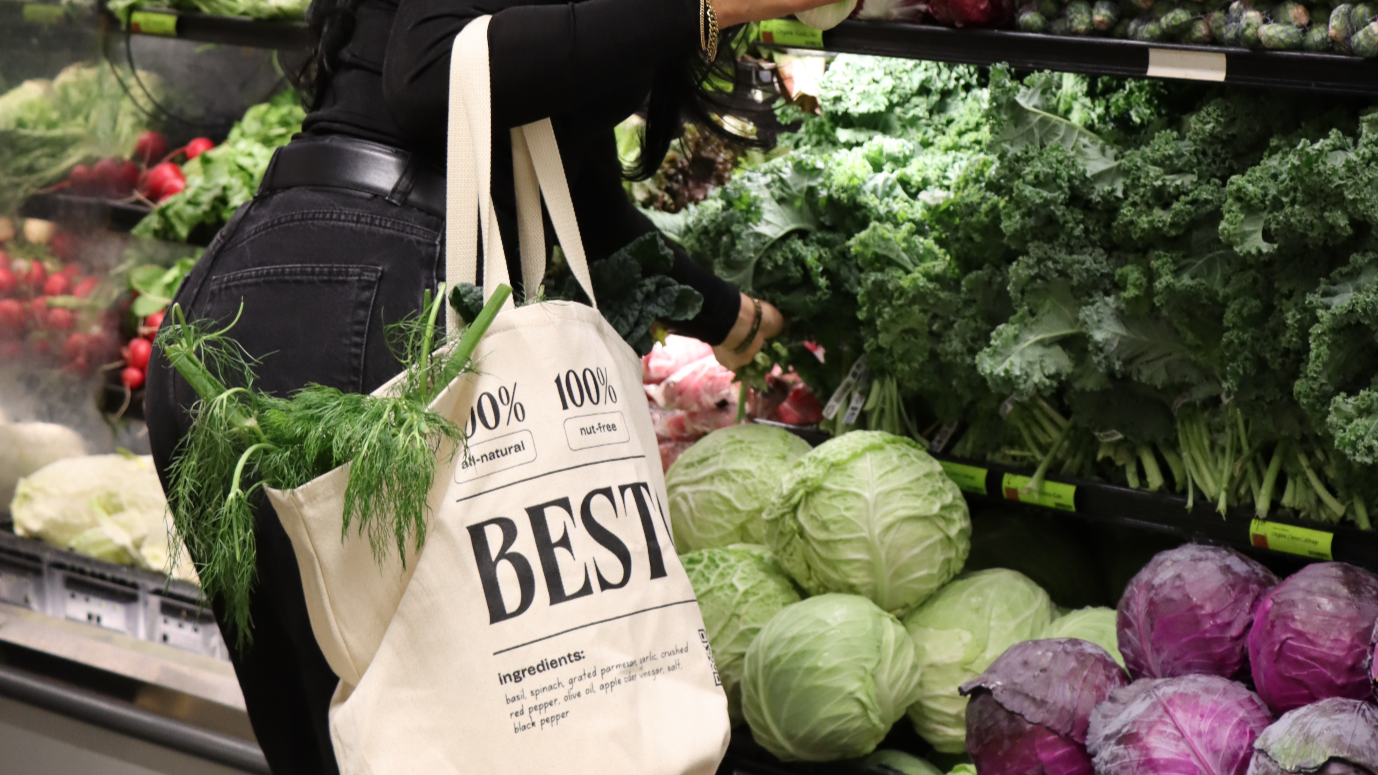 A woman holding a reusable tote bag filled with fresh vegetables, including broccoli kale, cabbage, celery, and dill, in a grocery stores produce section at Whole Foods. The bag displays text about Besto, natural and nut-free pesto.