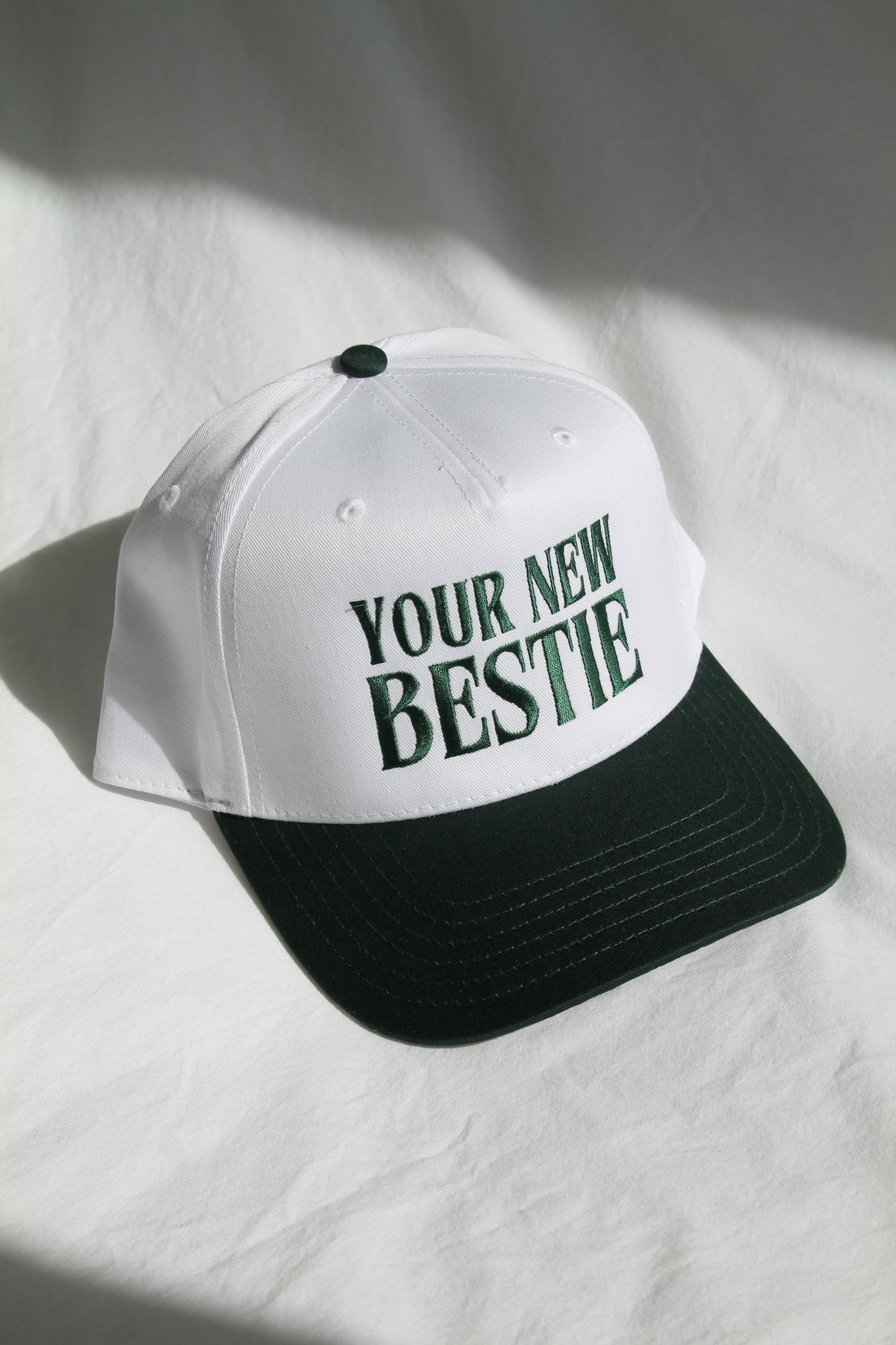 A white trucker hat has embroidered dark green letters "YOUR NEW BESTIE" with a dark visor. The hat is sitting on a white comforter with natural lighting and shadows.