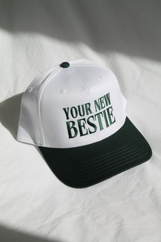 A white trucker hat has embroidered dark green letters "YOUR NEW BESTIE" with a dark visor. The hat is sitting on a white comforter with natural lighting and shadows.