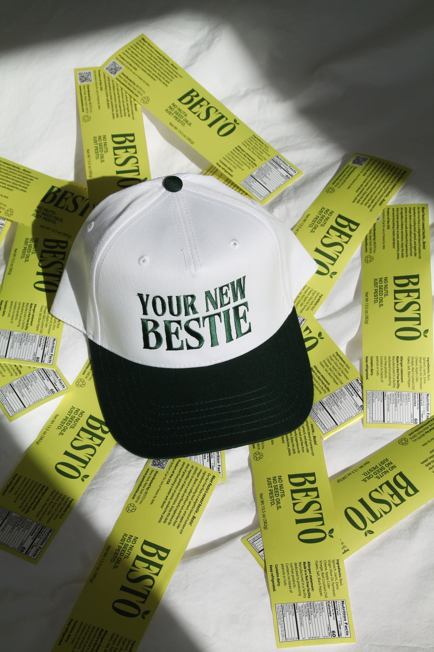 A white trucker hat with dark green letter embroidered stating "YOUR NEW BESTIE" with a dark green lid. The hat is laying on lime green Besto, natural nut free pesto labels on a white bed.