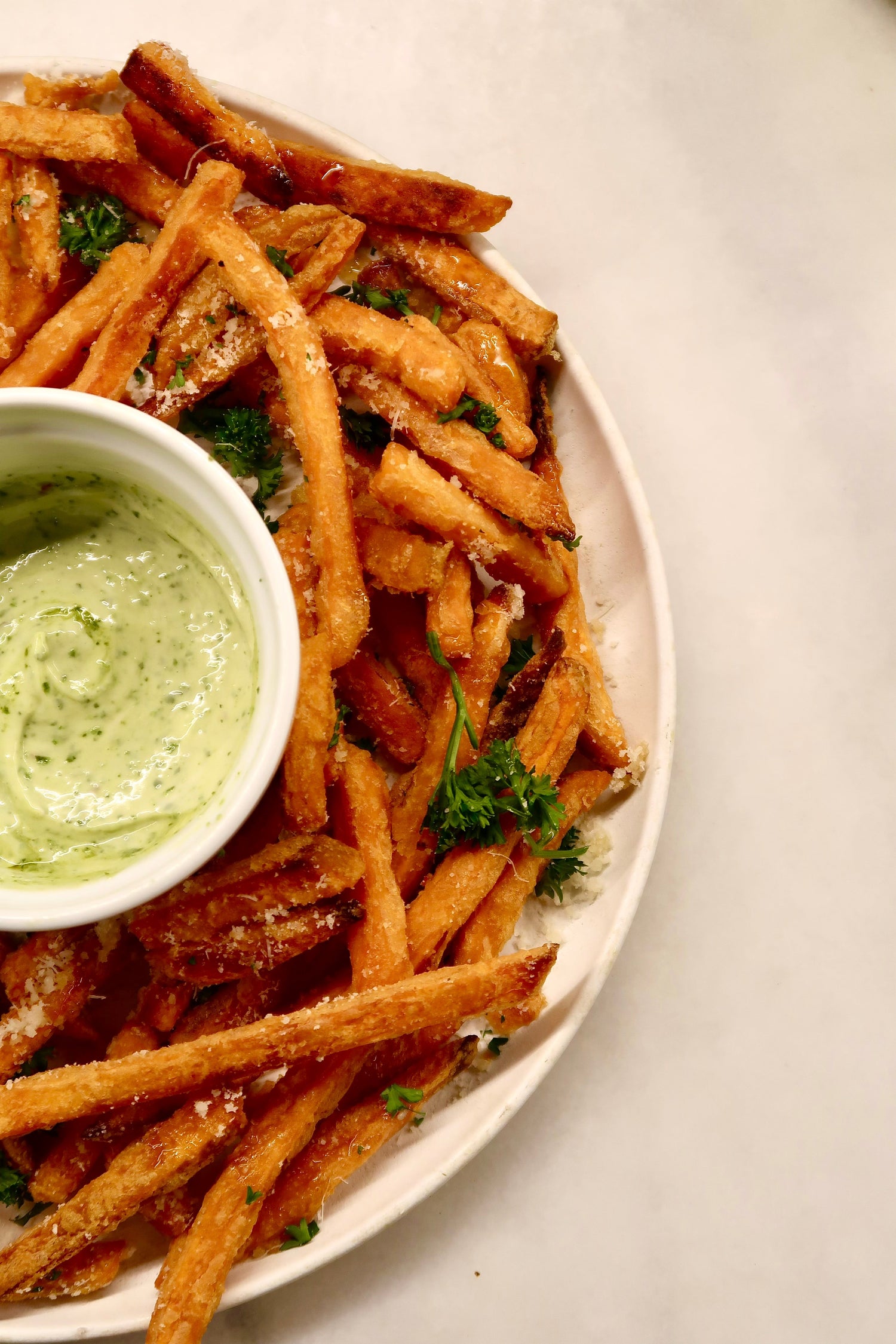 Homemade sweet potato fries on a white plate garnished with parmesan cheese and curly parsley. In the small, circular bowl is a pesto aioli made of Besto, mayo, Worcester sauce, and Dijon mustard.