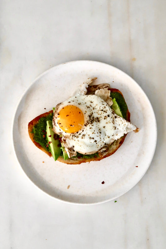 Pesto smeared on a slice of sourdough bread with avocado slices on top with a sunny side up egg sprinkled with black pepper and red crushed chili flakes served on white a plate.