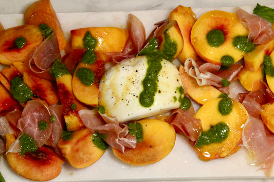 Peach slices on a platter with burrata in the center and pieces of prosciutto ham. Drizzled on top with nut free Besto pesto, honey, and olive oil with salt and black pepper. Delicious tapas and hosting appetizer.