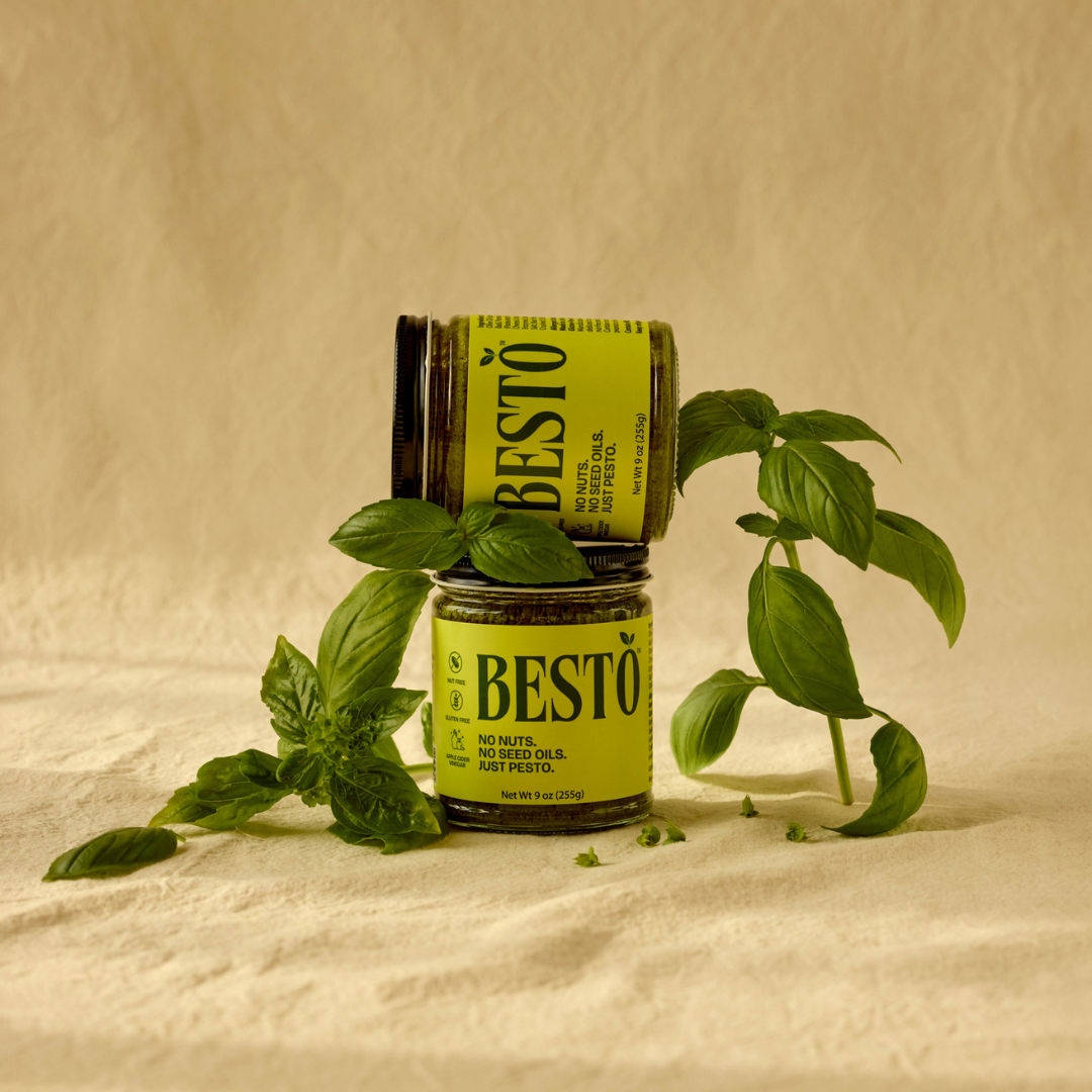 two jars of Besto Pesto with fresh basil leaves stacked on eachother beige background