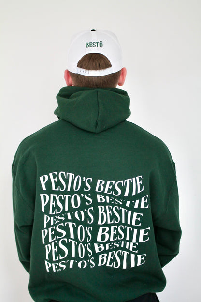 A boy is turned around against a white wall wearing a white trucker hat that has the logo "BESTO" with a leaf on the "O". He is wearing an oversized forest green hoodie with cool font on the back "Pesto's Bestie".