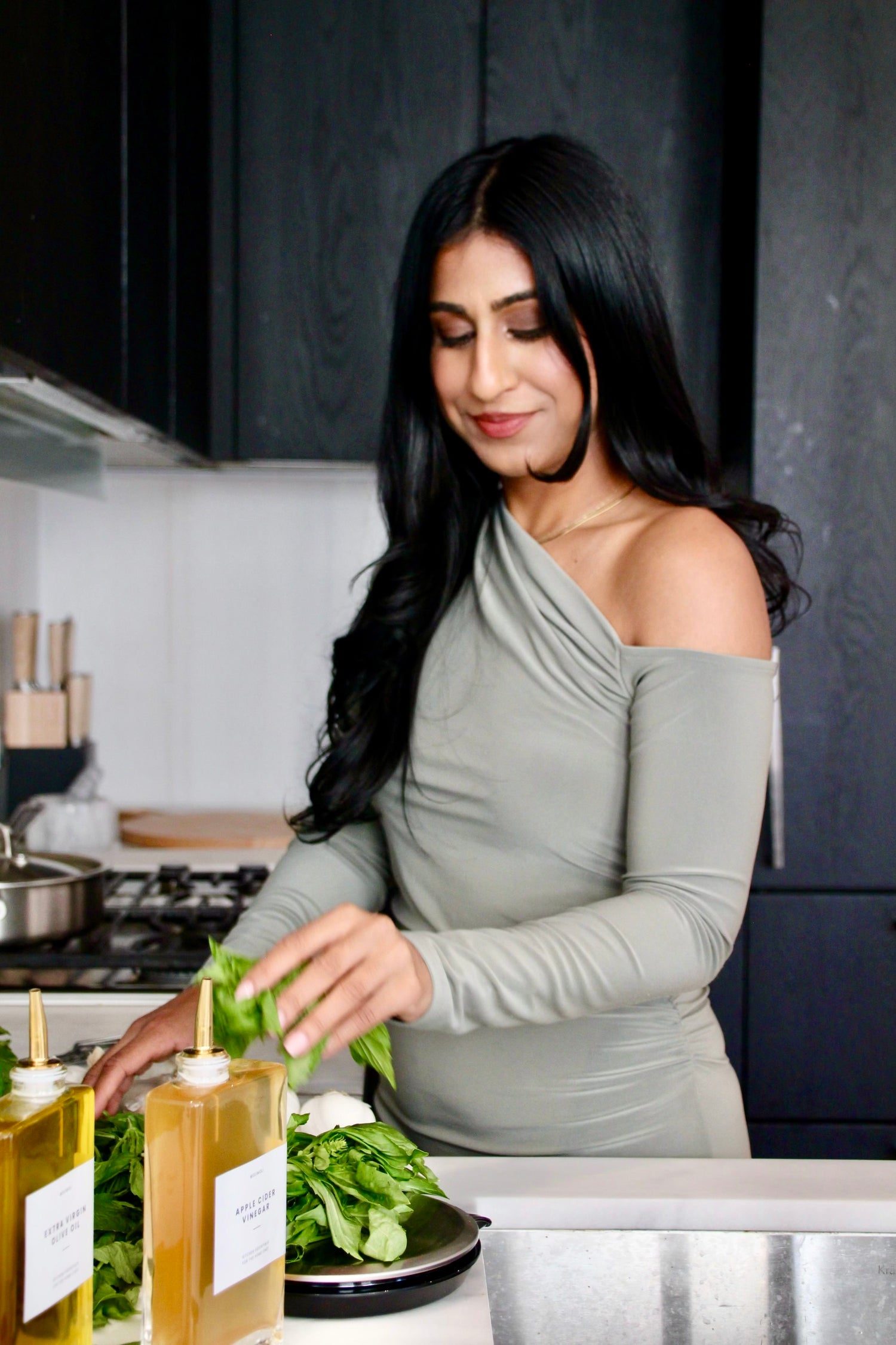 A woman with long black hair stands in a kitchen, smiling slightly while preparing a meal. She wears a light gray, off-shoulder dress and places green, basil leaves on a cutting board beside bottles of extra virgin oil and apple cider vinegar. The setting is modern with dark cabinets and modern interior design. 