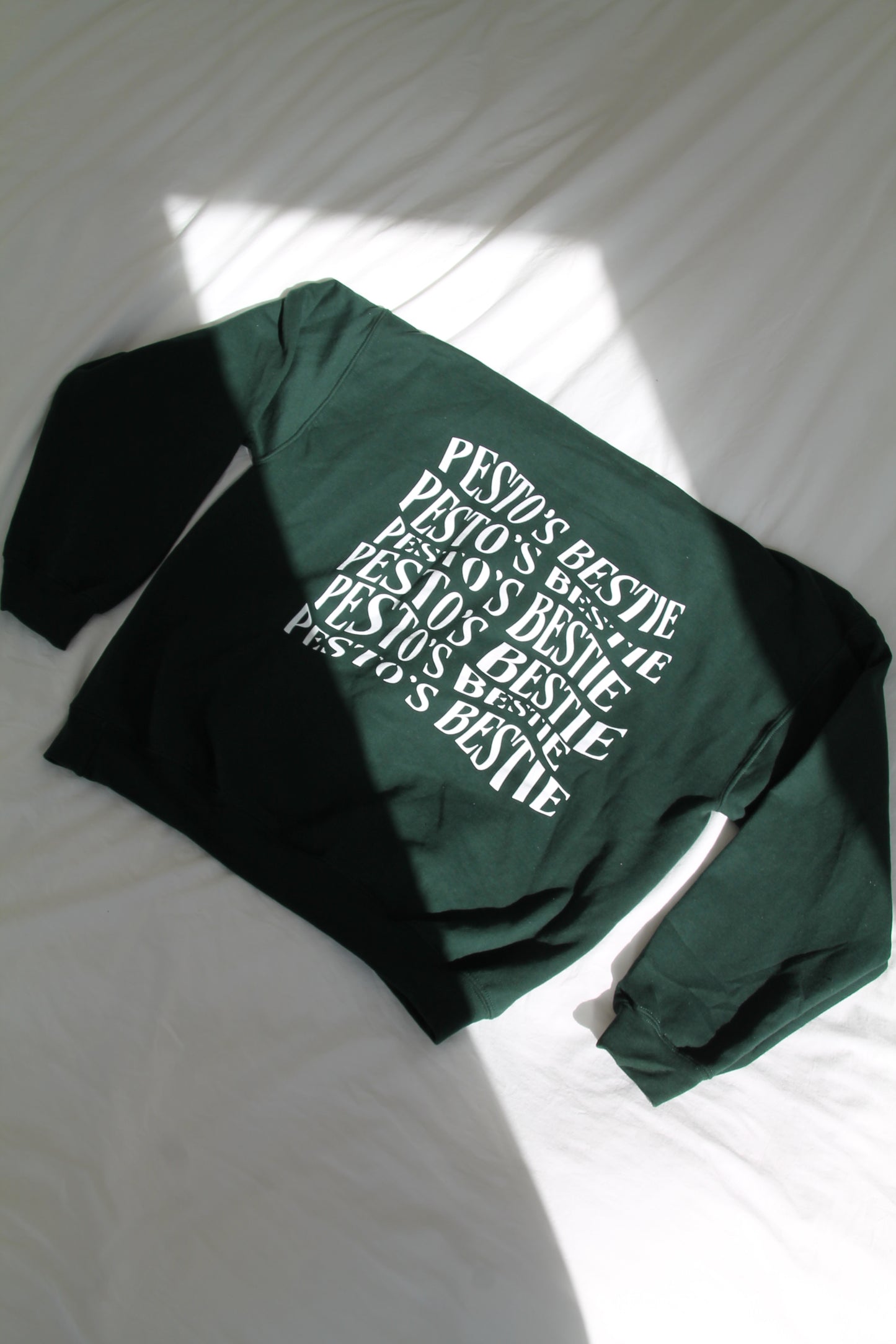 A dark forest green hoodie laid across a white modern bed. The back of the hoodie has in artistic font "Pesto's Bestie".