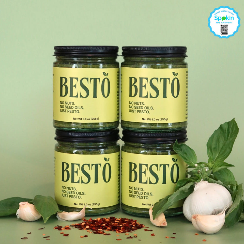 Four jars of Besto pesto stacked on top of each other against a green background. The lids are black and they have a bright green label stating "BESTO" with a leaf on the "O". Underneath the logo, the slogan "No Nuts. No Seed Oils. Just Pesto." The jars are 9 oz and 255 grams. Scattered below the jars are fresh basil leaves, garlic bulbs, and red crushed chili flakes.