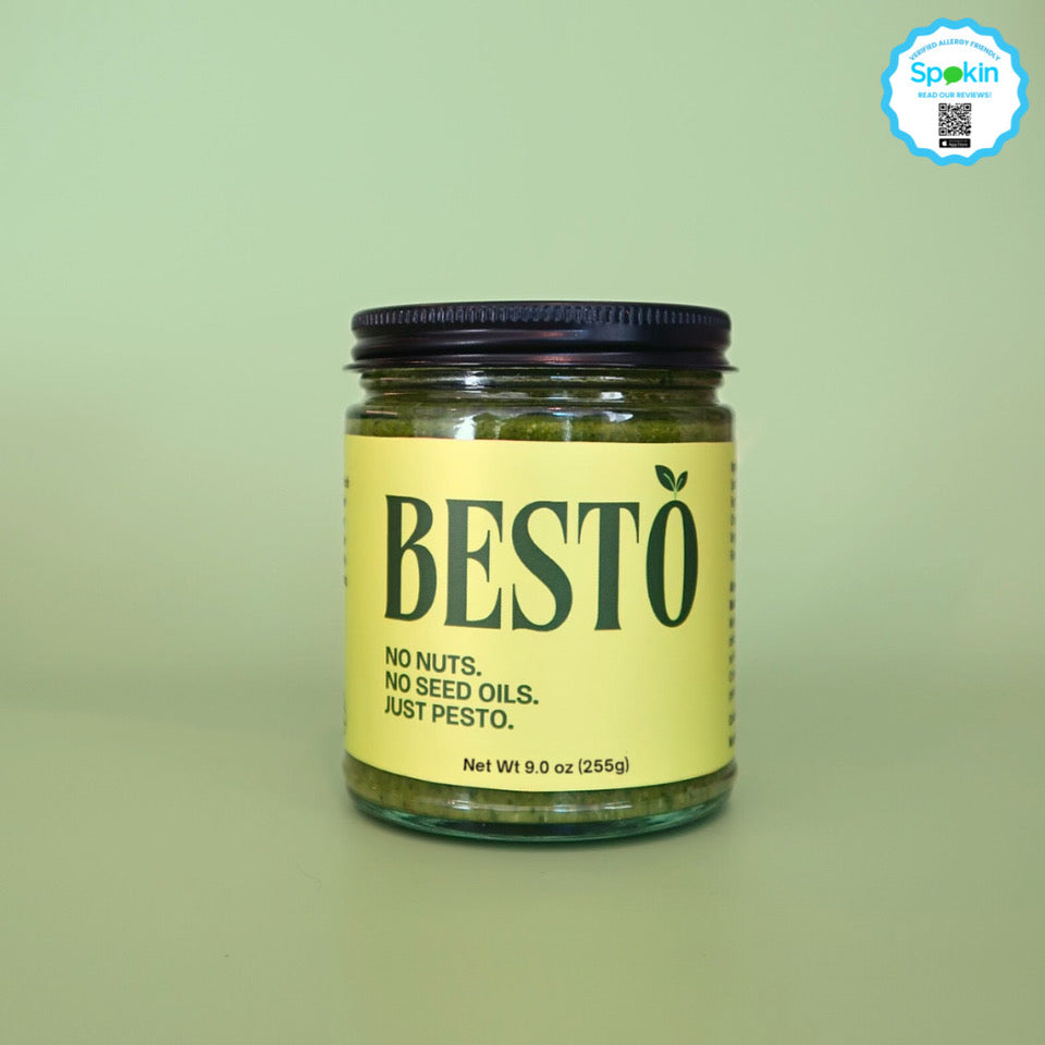 A 9oz jar of Besto pesto against a green background. The lid is black and on the glass jar is a bright green label with dark green letters "BESTO" with a green leaf on the "O". Underneath the logo is "No Nuts. No Seed Oils. Just Pesto."