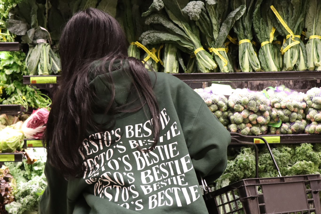 A woman in a dark, forest green hoodie that has trippy font saying "Pesto's Bestie" which is for the brand Besto, a nut free pesto. She is shopping at a green, vegetable produce section at Whole Foods grocery store. 