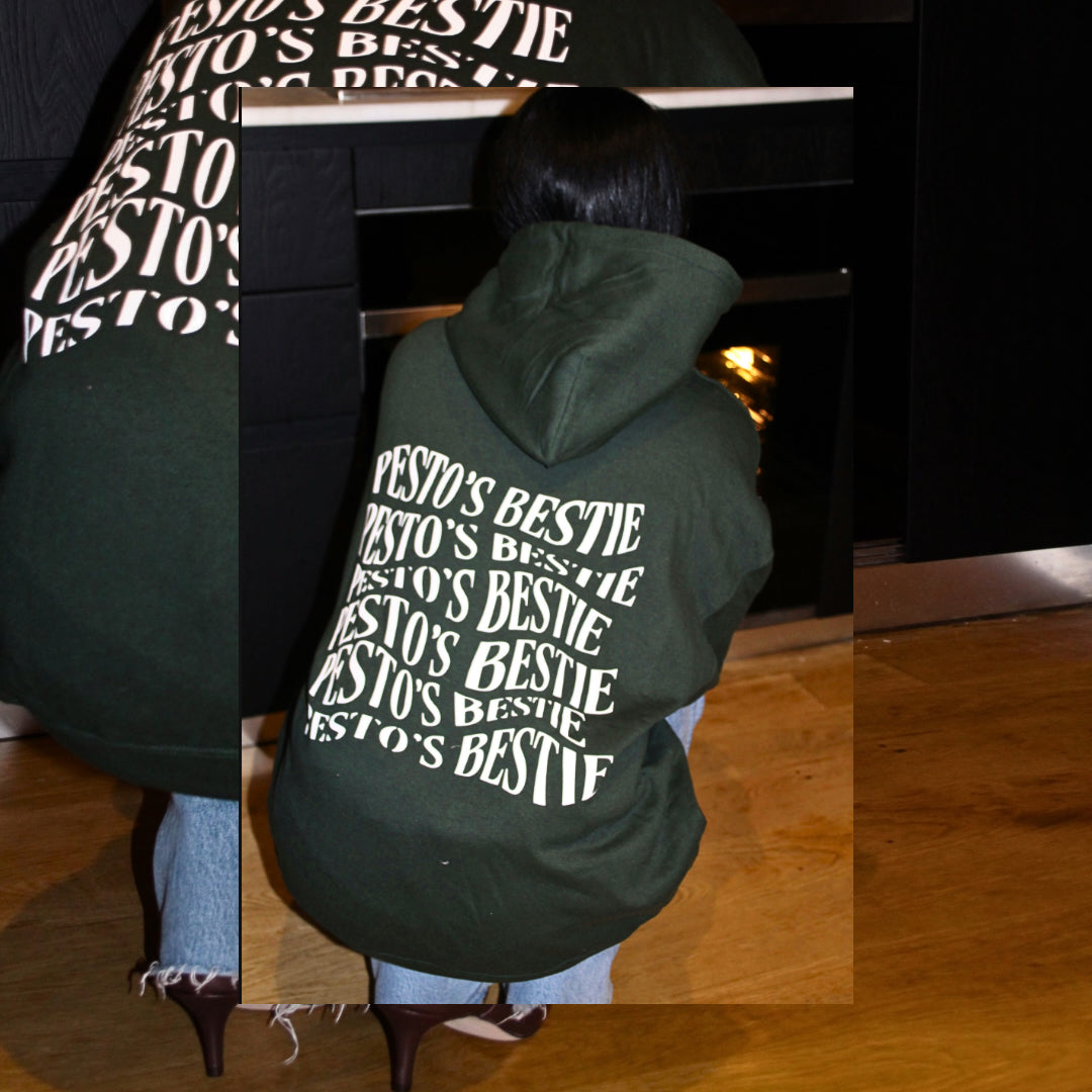 An artistic overlapped picture of a woman crouching down wearing a forest green hoodie with font "Pesto's bestie" in white letters. She is looking into a oven, wearing jeans, and high heeled brown boots and standing on hardwood flooring.