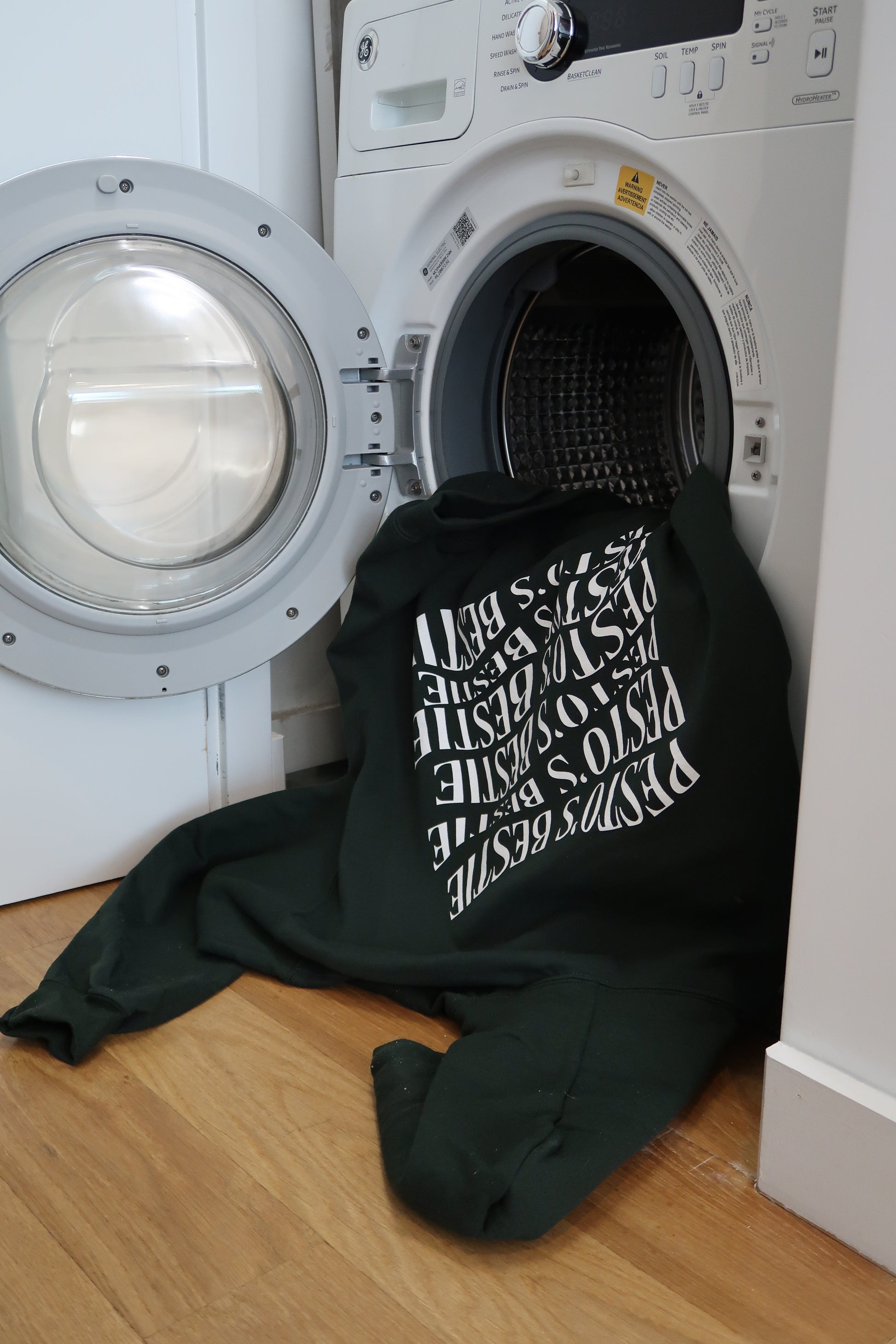 A forest green hoodie with white lettering on the back stating "Pesto's Bestie" is hanging out of the dryer or washing machine and touching the hardwood flooring. 