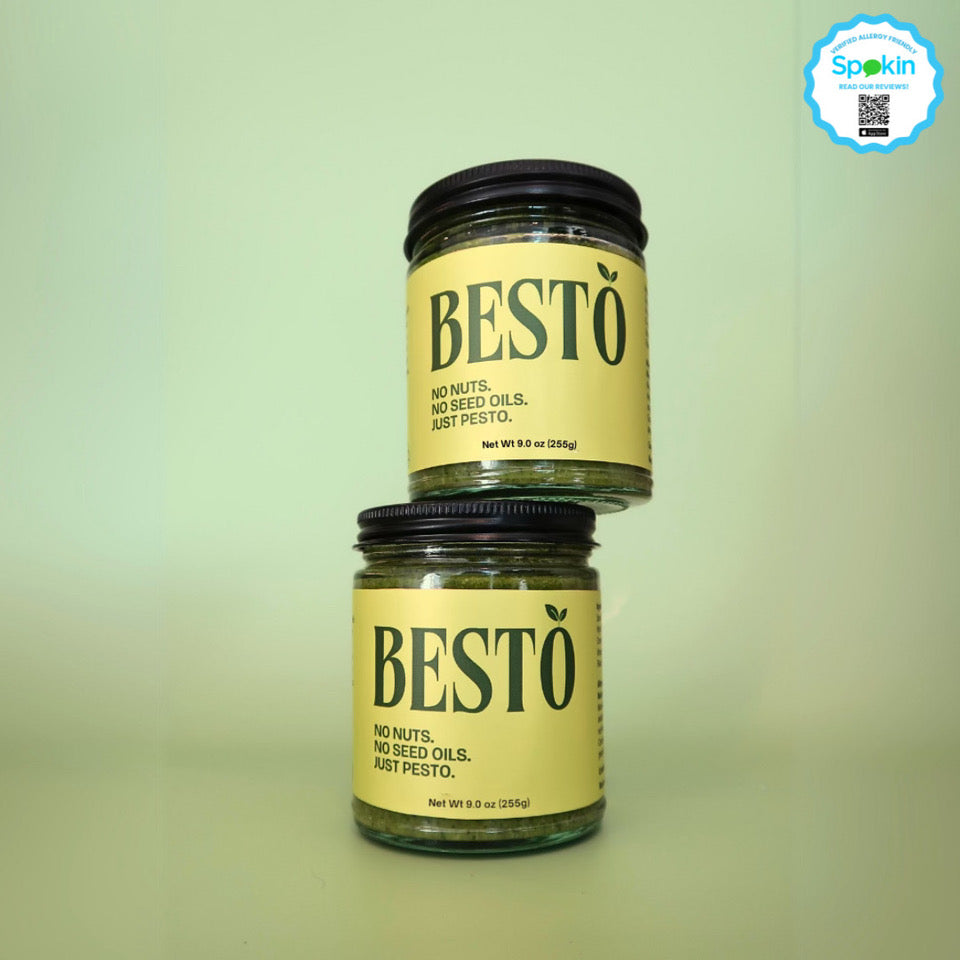 Two jars of Besto pesto stacked on top of each other against a green background. The lids are black and they have a bright green label stating "BESTO" with a leaf on the "O". Underneath the logo, the slogan "No Nuts. No Seed Oils. Just Pesto." The jars are 9 oz and 255 grams.