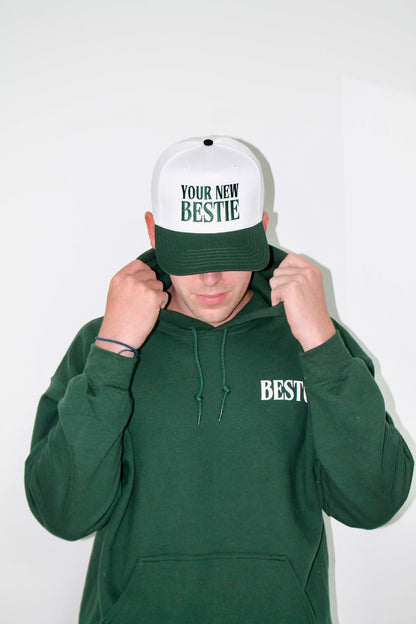 A boy wearing a white trucker hat embroidered with green letters "Your New Bestie" with a dark green visor. He is wearing a forest green hoodie with a logo on the top right chest in white letters "BESTO" with a leaf on the O. He is against a white wall. 